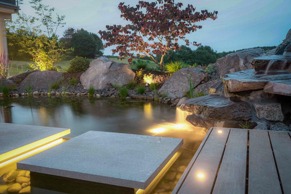 Teichanlage mit schwebenden Trittplatten und seitwärtigem Wasserfall in Abendstimmung